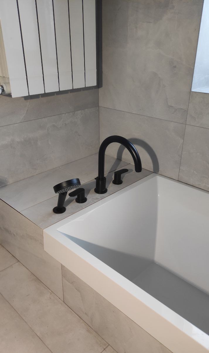 Nowoczesna bateria wannowa w kolorze czarnym, zamontowana na obudowie wanny z szarych płytek, z widocznym fragmentem grzejnika panelowego na ścianie.