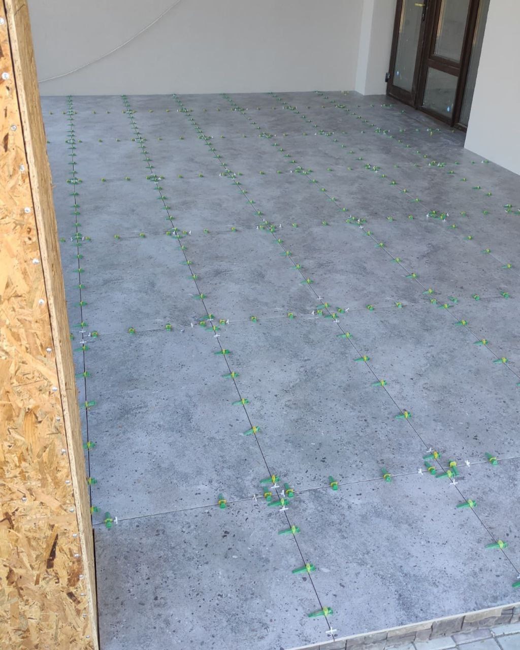 Układanie szarych płytek podłogowych z użyciem zielonych krzyżyków dystansowych, widoczne fragmenty drzwi i ściany.