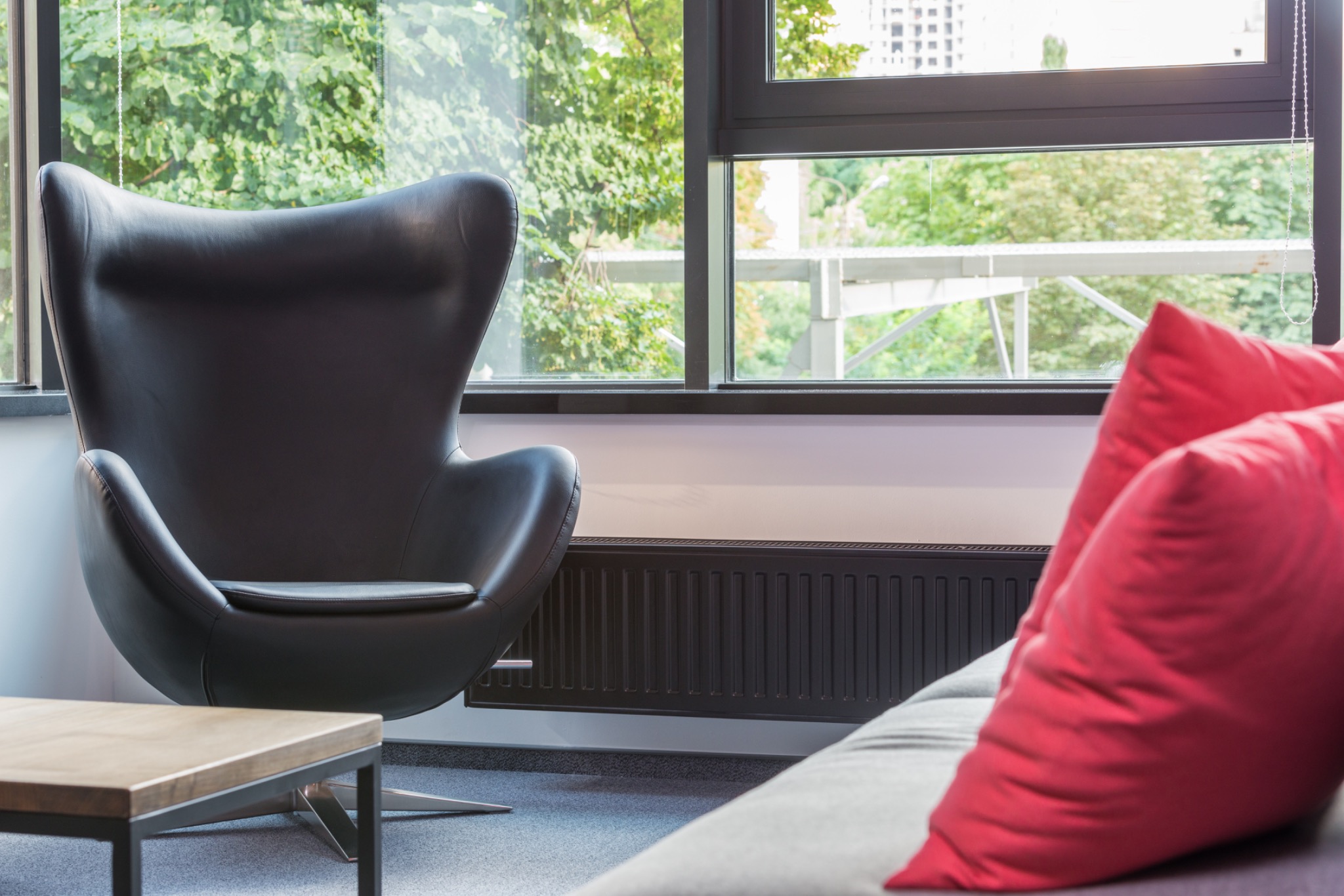 Nowoczesne wnętrze z czarnym fotelem Egg, widokiem na zieleń za oknem i czerwonymi poduszkami na szarej sofie.