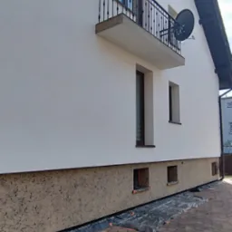 Dwukolorowa elewacja domu z balkonem i anteną satelitarną, widoczne okna z brązowymi ramami i zabezpieczenie folią fundamentów podczas prac wykończeniowych.