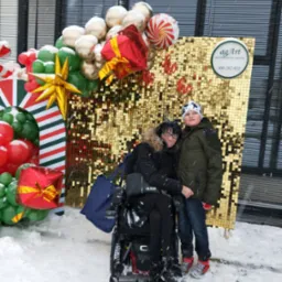 Dwie osoby pozują na tle złotej ściany z cekin&oacute;w i świątecznej dekoracji balonowej na zewnątrz, podczas śnieżnej pogody; widoczny logotyp firmy dekoratorskiej.