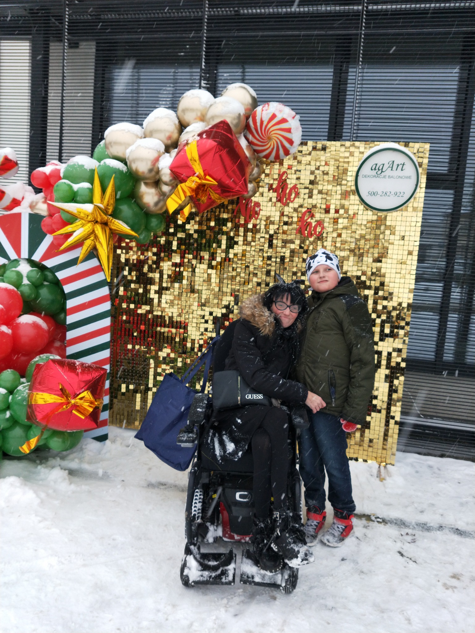 Dwie osoby pozują na tle złotej ściany z cekinów i świątecznej dekoracji balonowej na zewnątrz, podczas śnieżnej pogody; widoczny logotyp firmy dekoratorskiej.