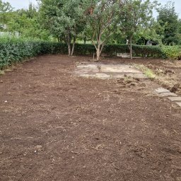 Czysty Dom - Przygotowany pod trawnik teren z resztkami trawy i płytami chodnikowymi, pomarańczowa łopata wbita w ziemię, żywopłot w tle.