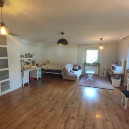 Czysty Dom - Przestronny pokój dzienny z drewnianą podłogą, białymi meblami, łóżkiem, sofą i oknami z roślinami, oświetlony trzema lampami sufitowymi.