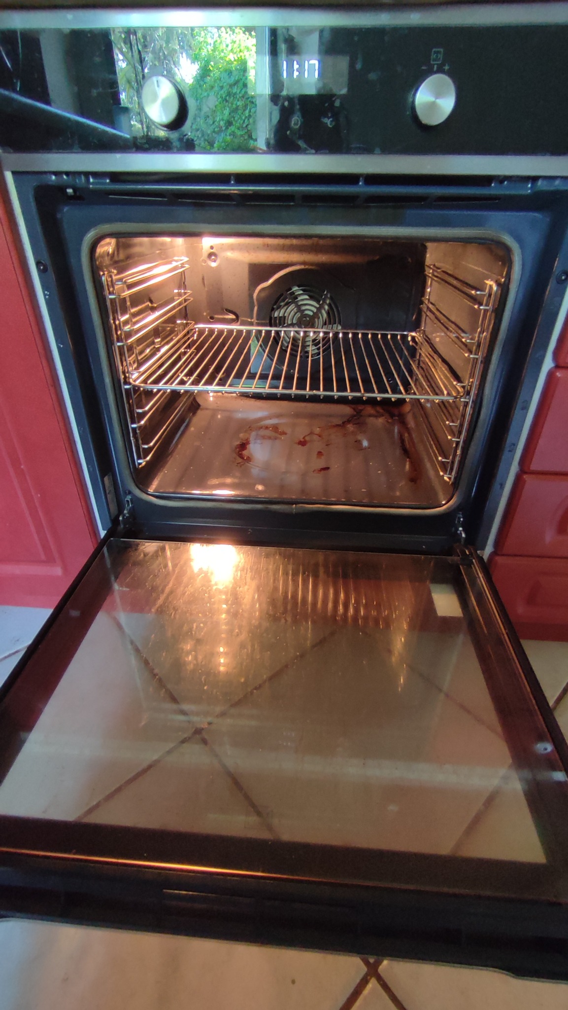 Wnętrze brudnego piekarnika z otwartymi drzwiczkami, widoczne resztki jedzenia na dnie i tłuszcz na ściankach, metalowe ruszty wewnątrz, odbicie światła na szklanych drzwiach.