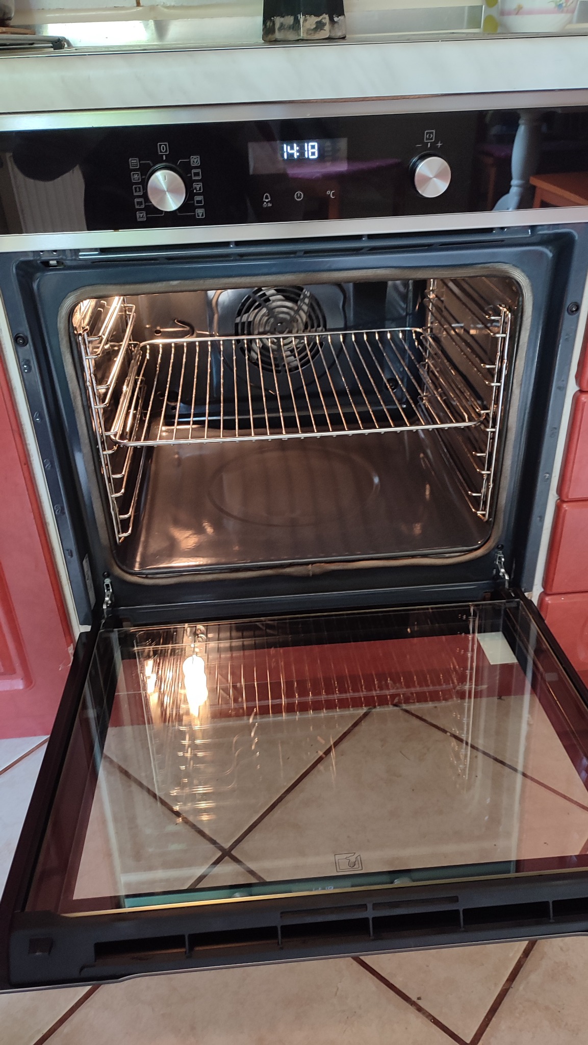 Otwarty piekarnik elektryczny z widocznymi rusztami, wentylatorem i odbiciem światła w szklanych drzwiach, ustawiony w kuchni z kafelkami na ścianie.