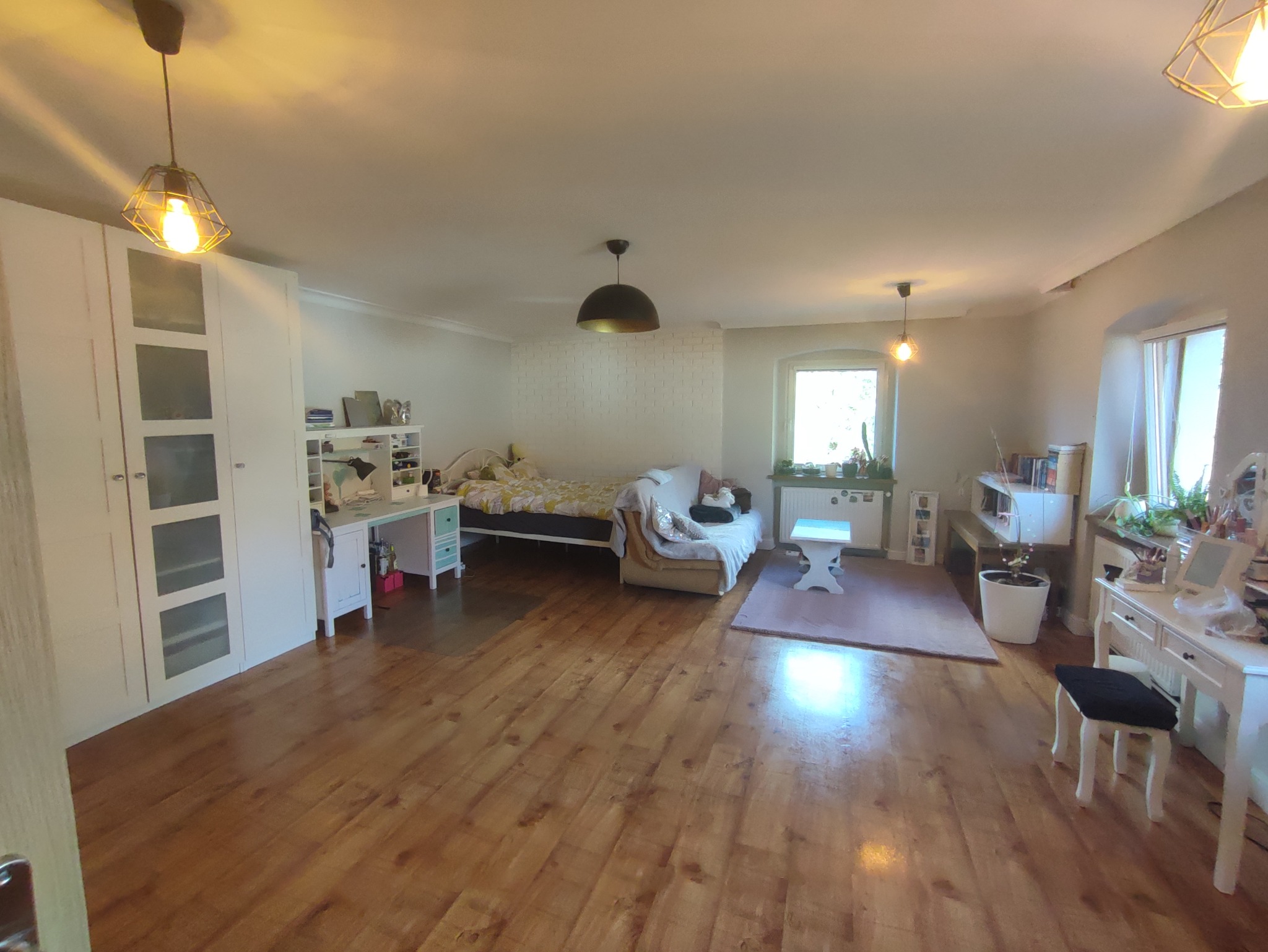 Przestronny pokój dzienny z drewnianą podłogą, białymi meblami, łóżkiem, sofą i oknami z roślinami, oświetlony trzema lampami sufitowymi.