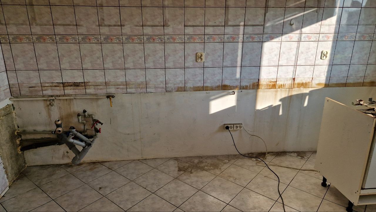 Pusta kuchnia w trakcie demontażu, widoczne odsłonięte rury, gniazdka elektryczne i ślady po starych płytkach na ścianach.