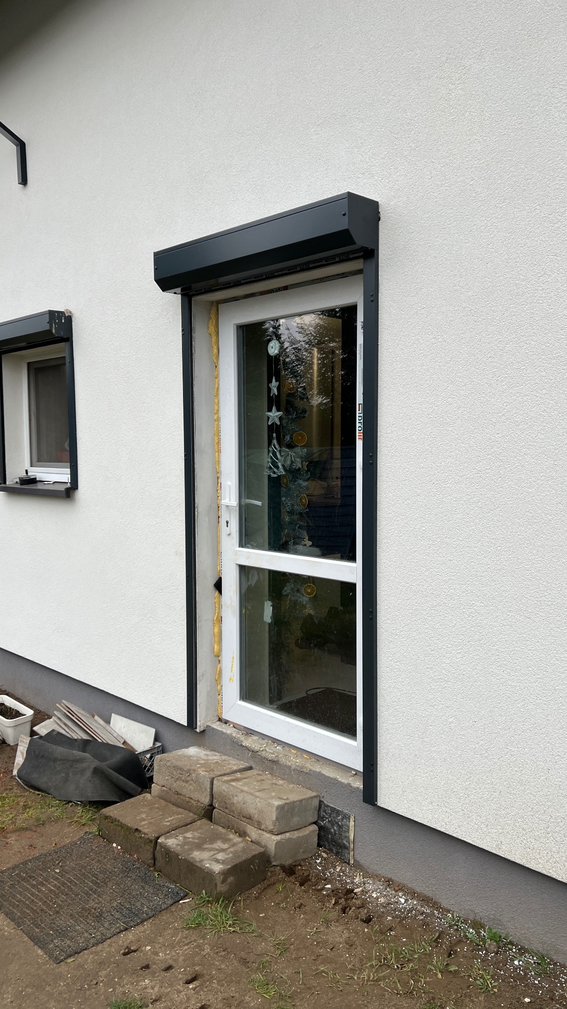 Montaż rolety zewnętrznej w oknie z widoczną pianką montażową i elementami wykończeniowymi. Na parapecie okna obok widoczna roleta. Elewacja budynku w jasnym kolorze.