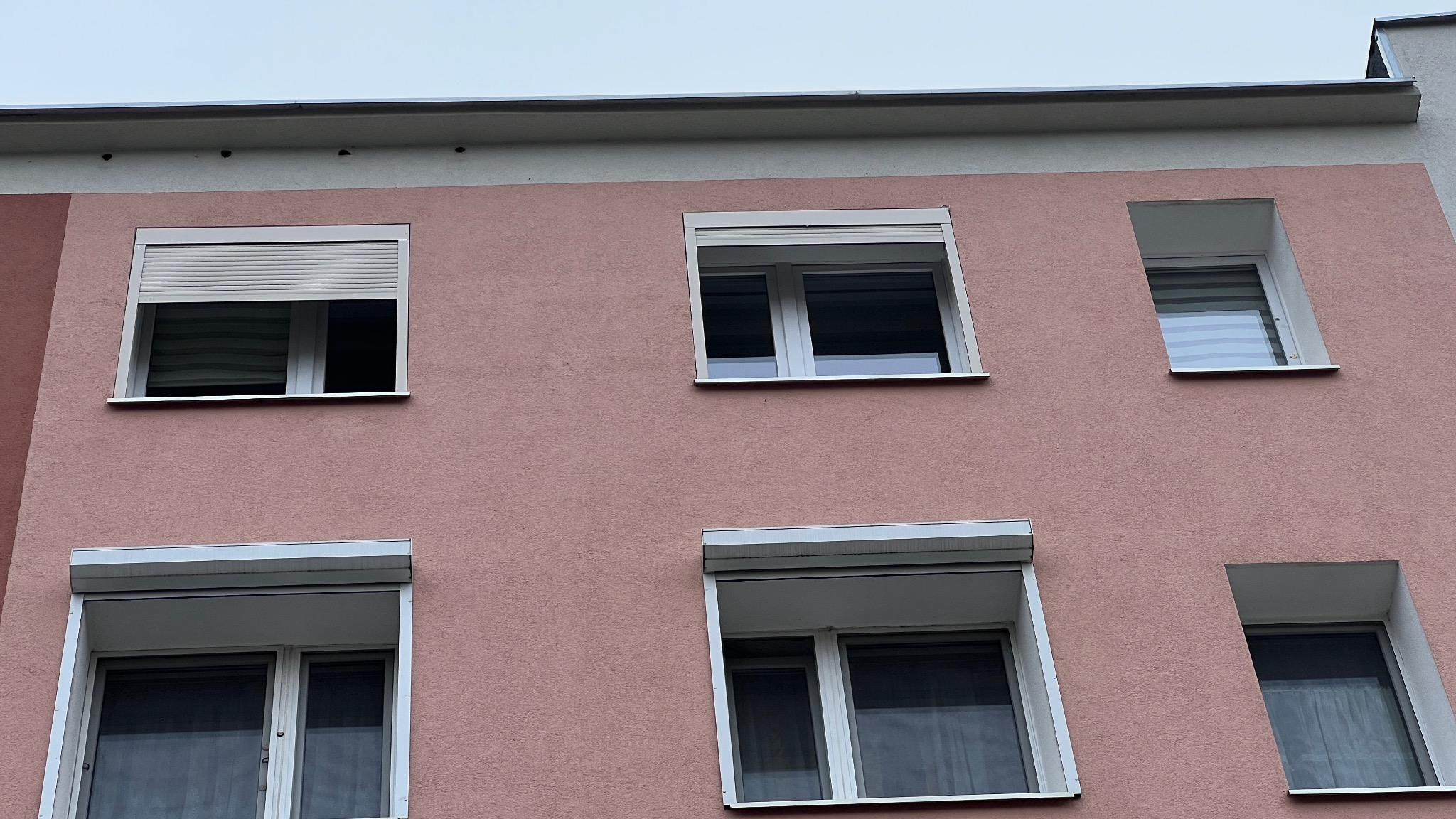 Elewacja budynku z oknami, niektóre z częściowo opuszczonymi roletami zewnętrznymi w kolorze białym. Różowa fasada z widoczną strukturą tynku.