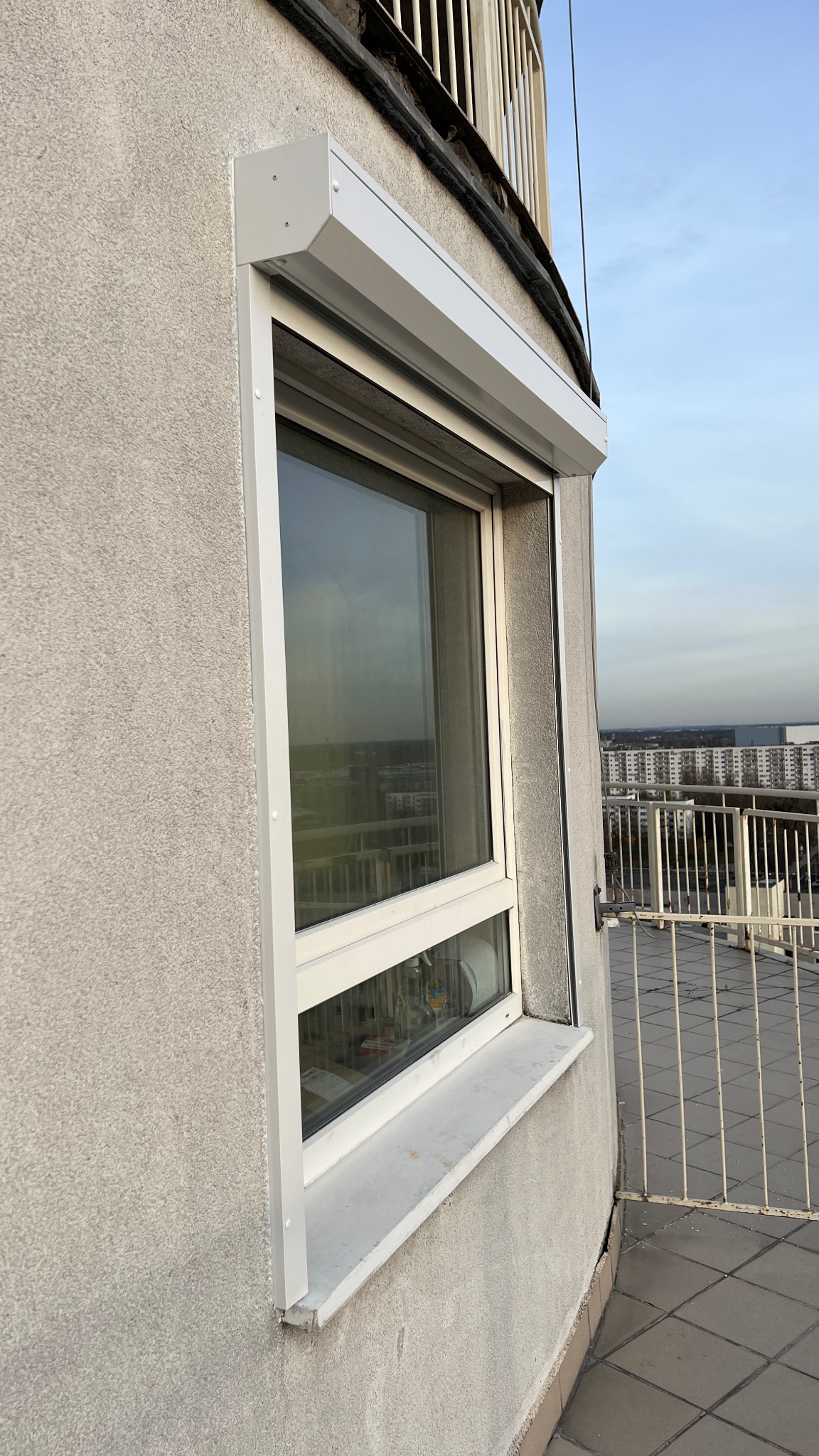 Zamknięta, biała roleta zewnętrzna na oknie budynku wielorodzinnego, widok z balkonu. Roleta zamontowana na oknie z widokiem na miasto w tle.