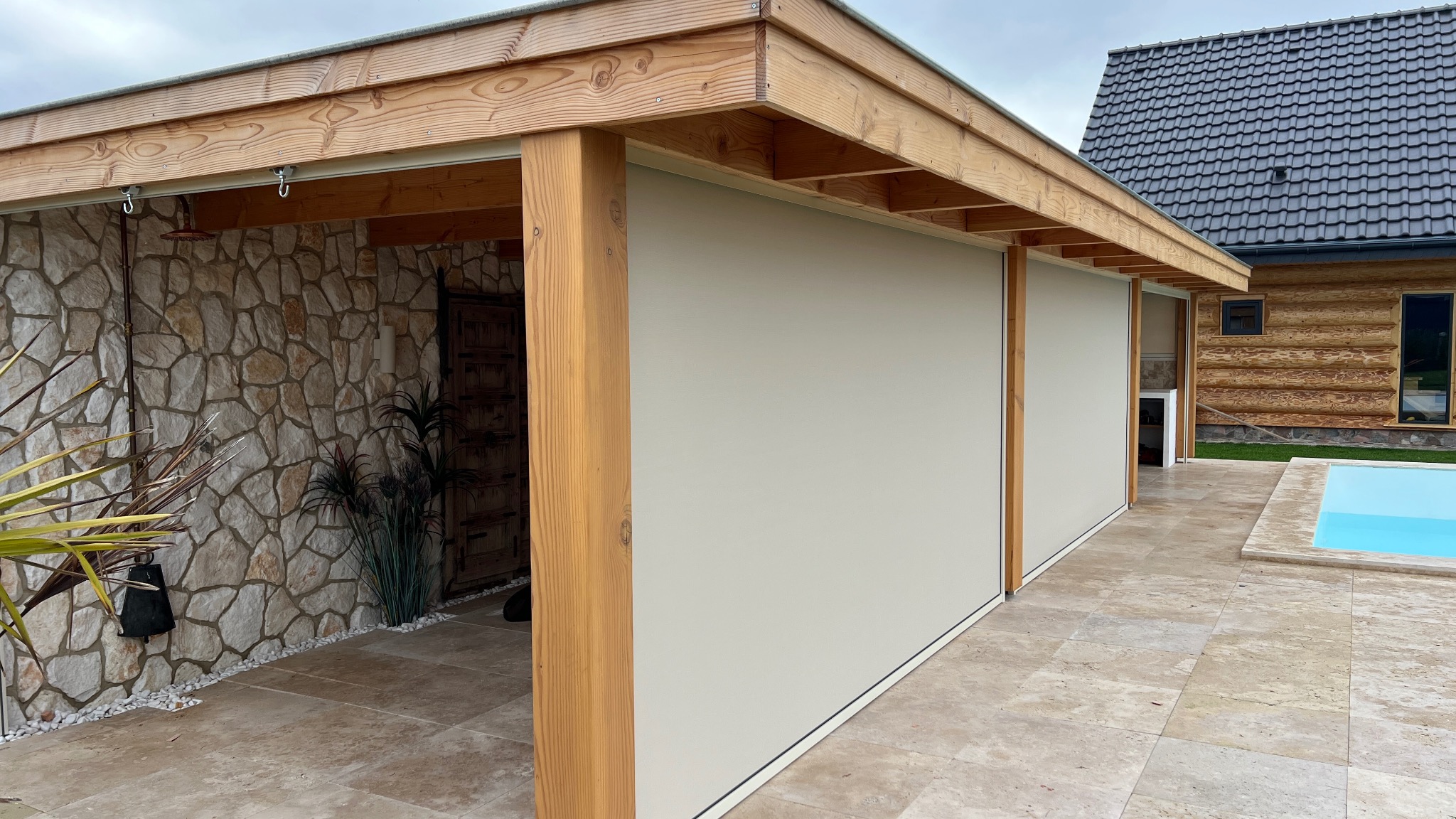 Beżowe rolety zewnętrzne w drewnianej konstrukcji altany, z kamienną ścianą w tle i basenem obok drewnianego domu. Widok na zewnątrz.