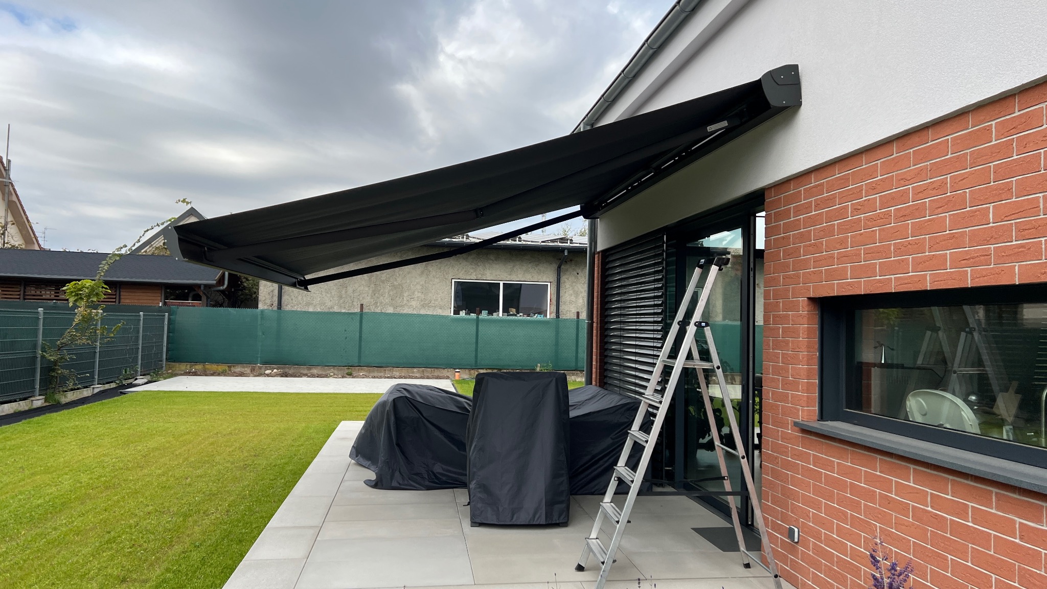 Rozłożona, czarna markiza tarasowa zamontowana nad oknem w nowoczesnym domu z ceglaną elewacją. Aluminiowa drabina oparta o ścianę. Widok na zadbany trawnik i meble ogrodowe.