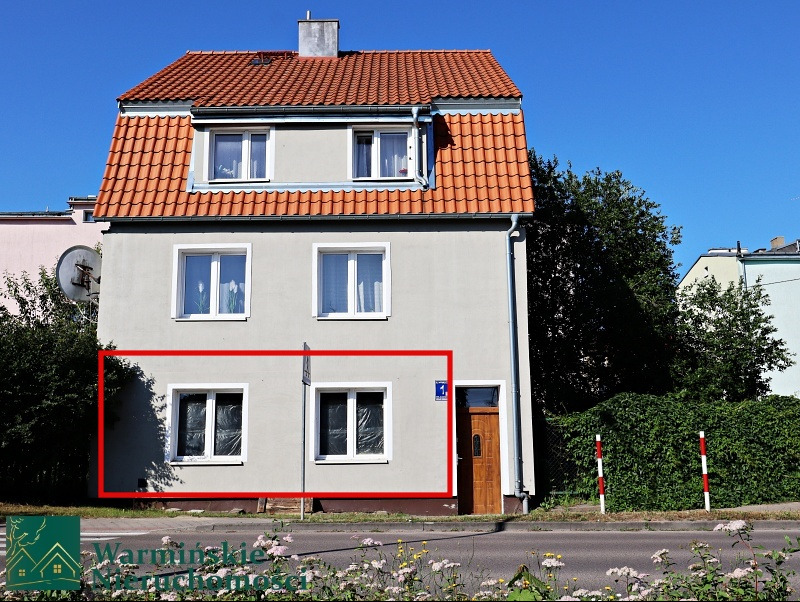 Dwupiętrowy dom z szarą elewacją i pomarańczowym dachem, z numerem domu '1' obok drewnianych drzwi wejściowych, widoczny z poziomu ulicy z kwiatami wzdłuż chodnika.