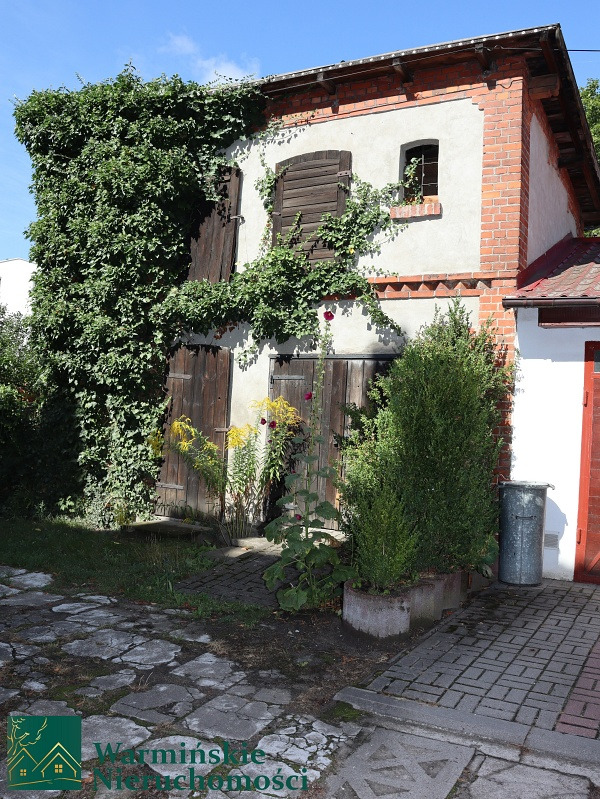 Dwukondygnacyjny budynek gospodarczy z ceglanymi narożnikami, częściowo pokryty bluszczem, z drewnianymi okiennicami i brukowanym podjazdem.