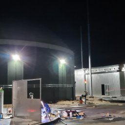 ARZO-SPAW - Nocna sceneria z pracami spawalniczymi przy konstrukcji metalowej, widoczny spawacz w kombinezonie, butla z gazem, spawarka i metalowa rama na tle oświetlonego zbiornika i budynku.