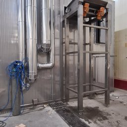 ARZO-SPAW - Metalowa konstrukcja z podajnikiem w hali produkcyjnej, obok rury wentylacyjne i niebieski przewód sprężonego powietrza, widoczna kratka ściekowa w podłodze.