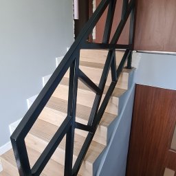 ARZO-SPAW - Nowoczesna, czarna, metalowa balustrada schodowa o geometrycznym wzorze, zamontowana na drewnianych schodach w jasnym wnętrzu.