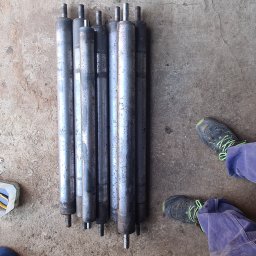 ARZO-SPAW - Rzędem ułożone, długie, metalowe wałki z wystającymi trzpieniami, leżące na betonowej posadzce, obok narzędzi i osoby w roboczych spodniach.