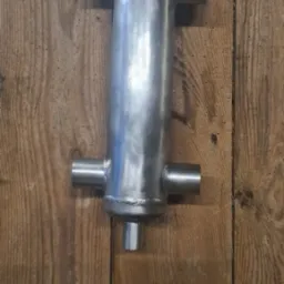 Surowy, spawany wymiennik ciepła ze stali nierdzewnej z czterema króćcami, zawieszony na tle drewnianej ściany.