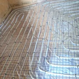 Rozłożone białe rury ogrzewania podłogowego na folii z nadrukiem w niebieską kratkę, przymocowane czarnymi klipsami, widok z góry pod kątem, surowe ściany w tle.