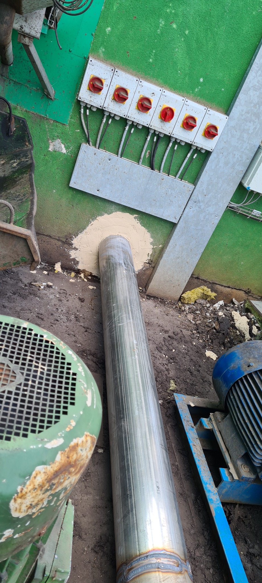 Metalowa rura przechodząca przez otwór w ścianie, uszczelniona zaprawą, z widocznymi spawami, obok fragmenty urządzeń mechanicznych i skrzynka z wyłącznikami.
