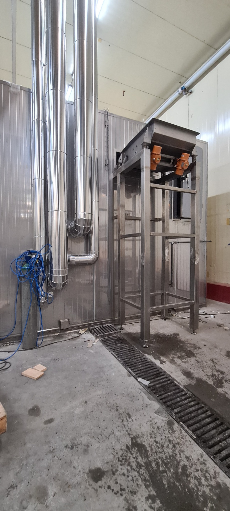 Metalowa konstrukcja z podajnikiem w hali produkcyjnej, obok rury wentylacyjne i niebieski przewód sprężonego powietrza, widoczna kratka ściekowa w podłodze.
