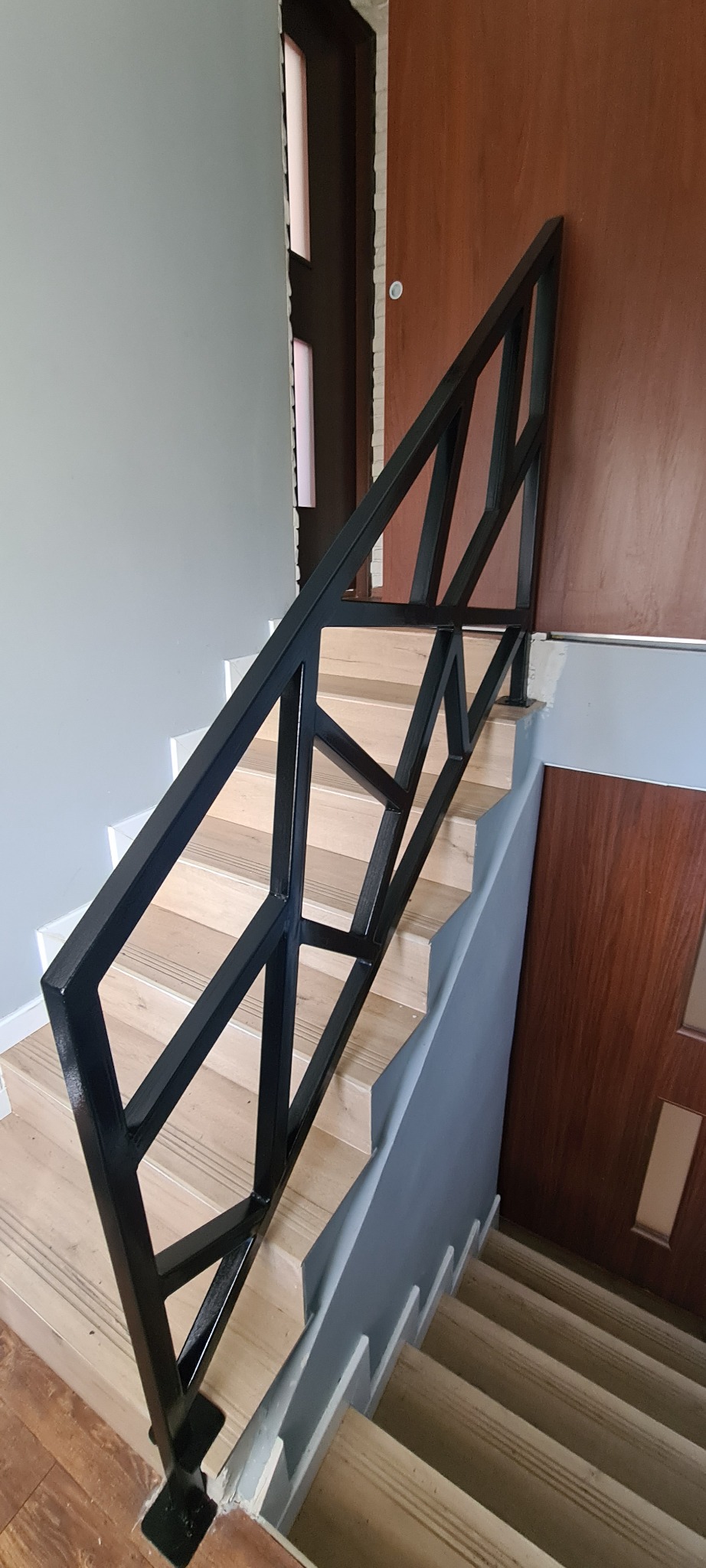 Nowoczesna, czarna, metalowa balustrada schodowa o geometrycznym wzorze, zamontowana na drewnianych schodach w jasnym wnętrzu.