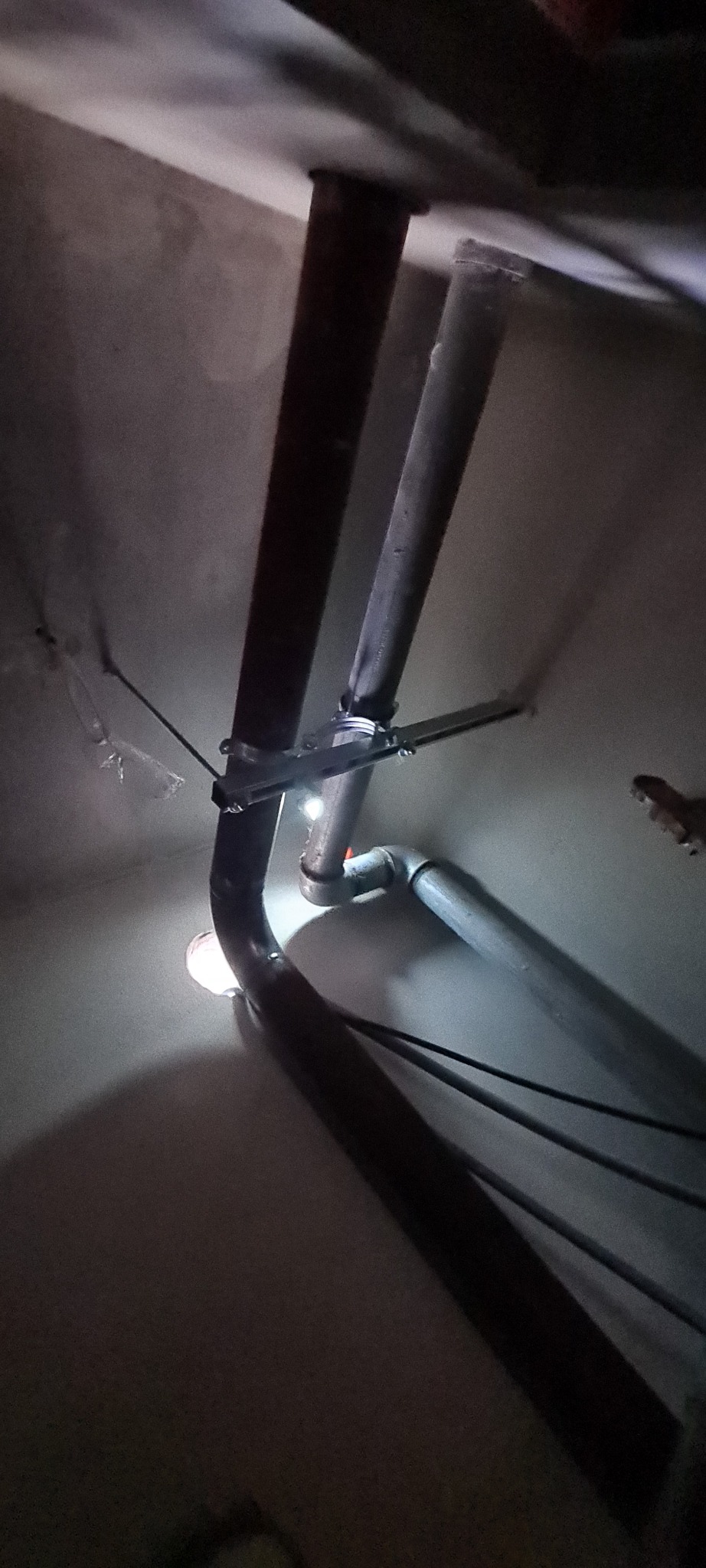 Rury kanalizacyjne pod sufitem, widoczne mocowania i łączenia, oświetlone punktowym światłem.