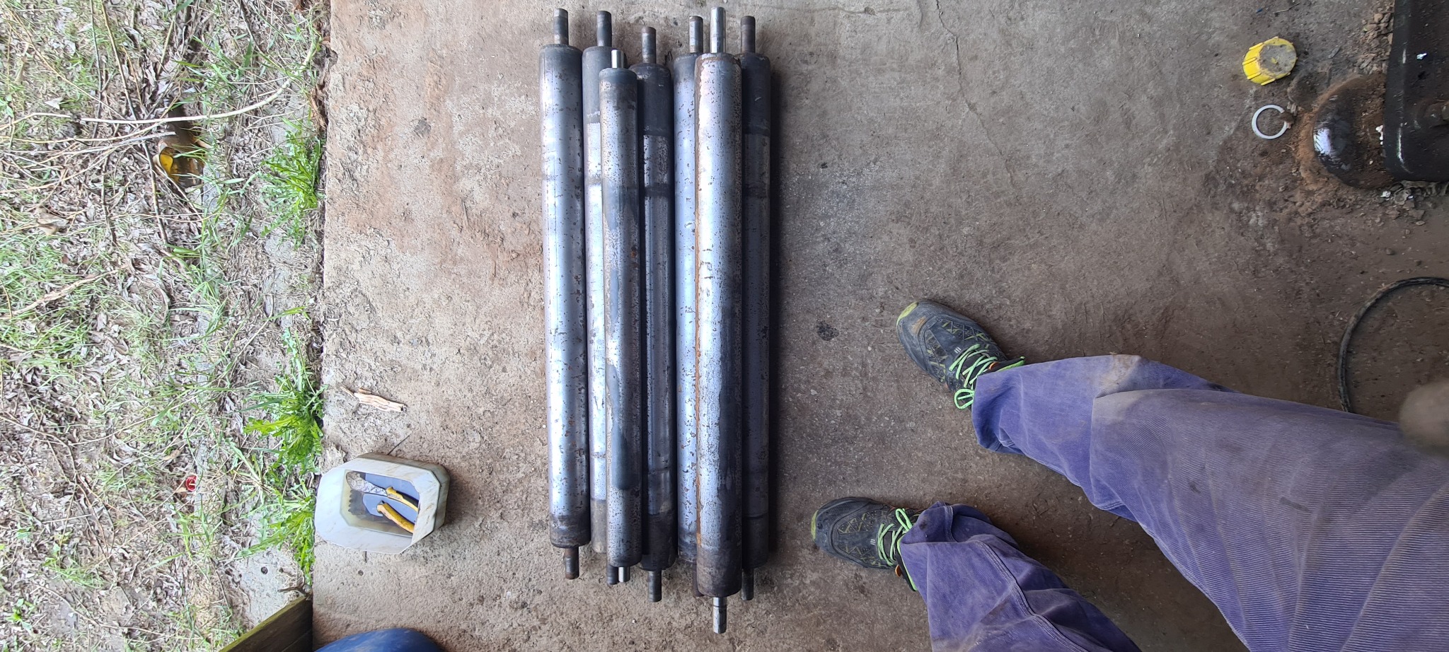 Rzędem ułożone, długie, metalowe wałki z wystającymi trzpieniami, leżące na betonowej posadzce, obok narzędzi i osoby w roboczych spodniach.