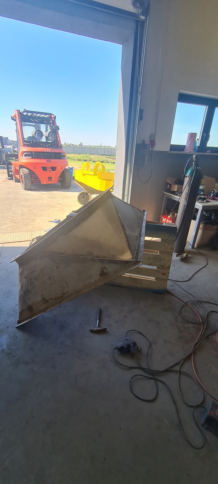 Metalowa konstrukcja na palecie w warsztacie, widoczne spawane łączenia, młotek i szlifierka, w tle wózek widłowy i żółta konstrukcja.