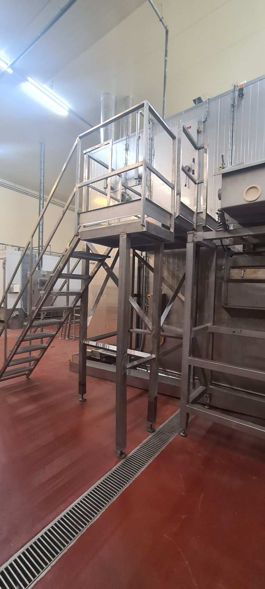 Metalowa konstrukcja platformy z schodami w hali przemysłowej, widoczne rury wentylacyjne i kratka ściekowa w posadzce.