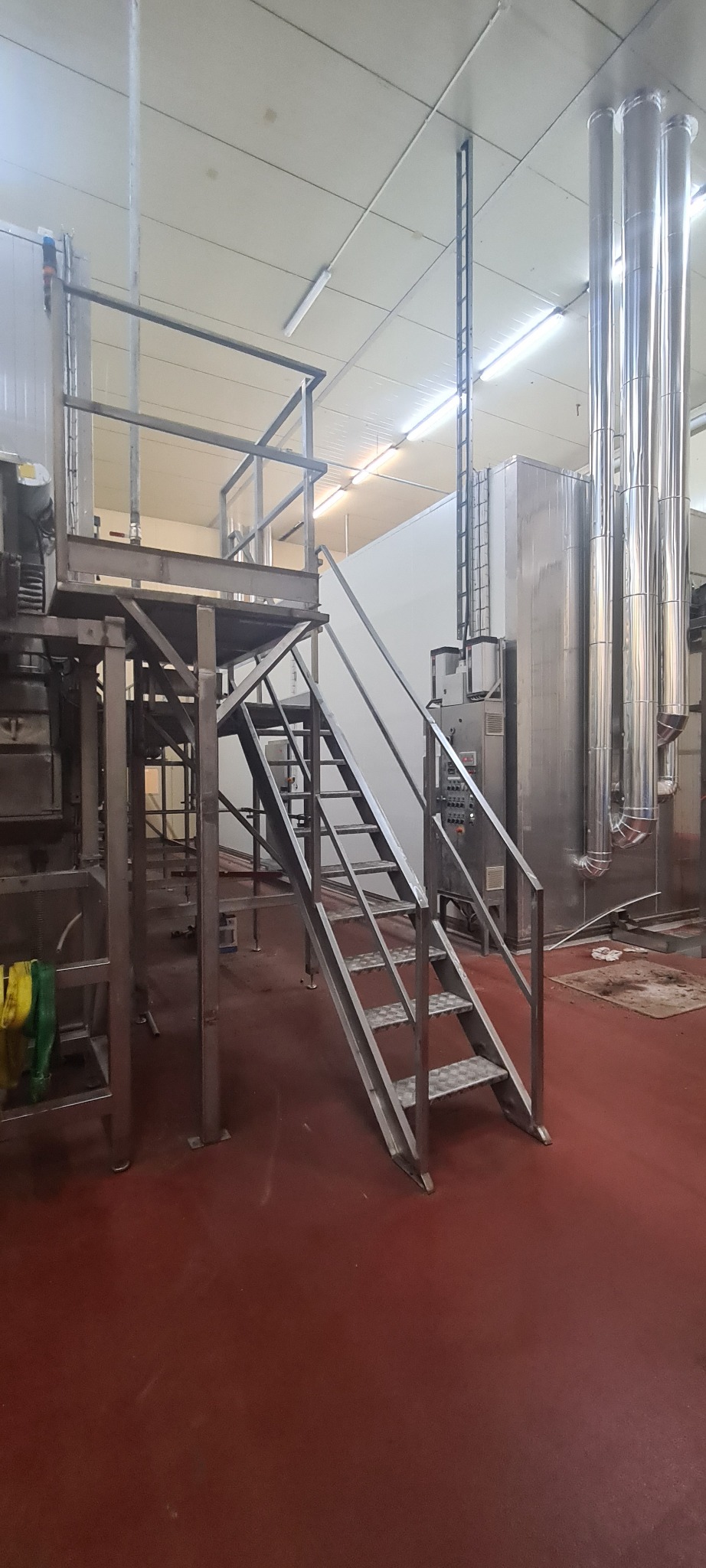 Metalowa konstrukcja schodów z platformą w hali przemysłowej, widoczne rury i maszyny w tle.