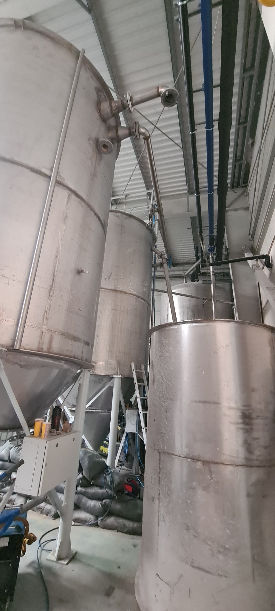 Metalowe zbiorniki przemysłowe w hali produkcyjnej, z widocznymi rurami, przewodami i workami z materiałem.