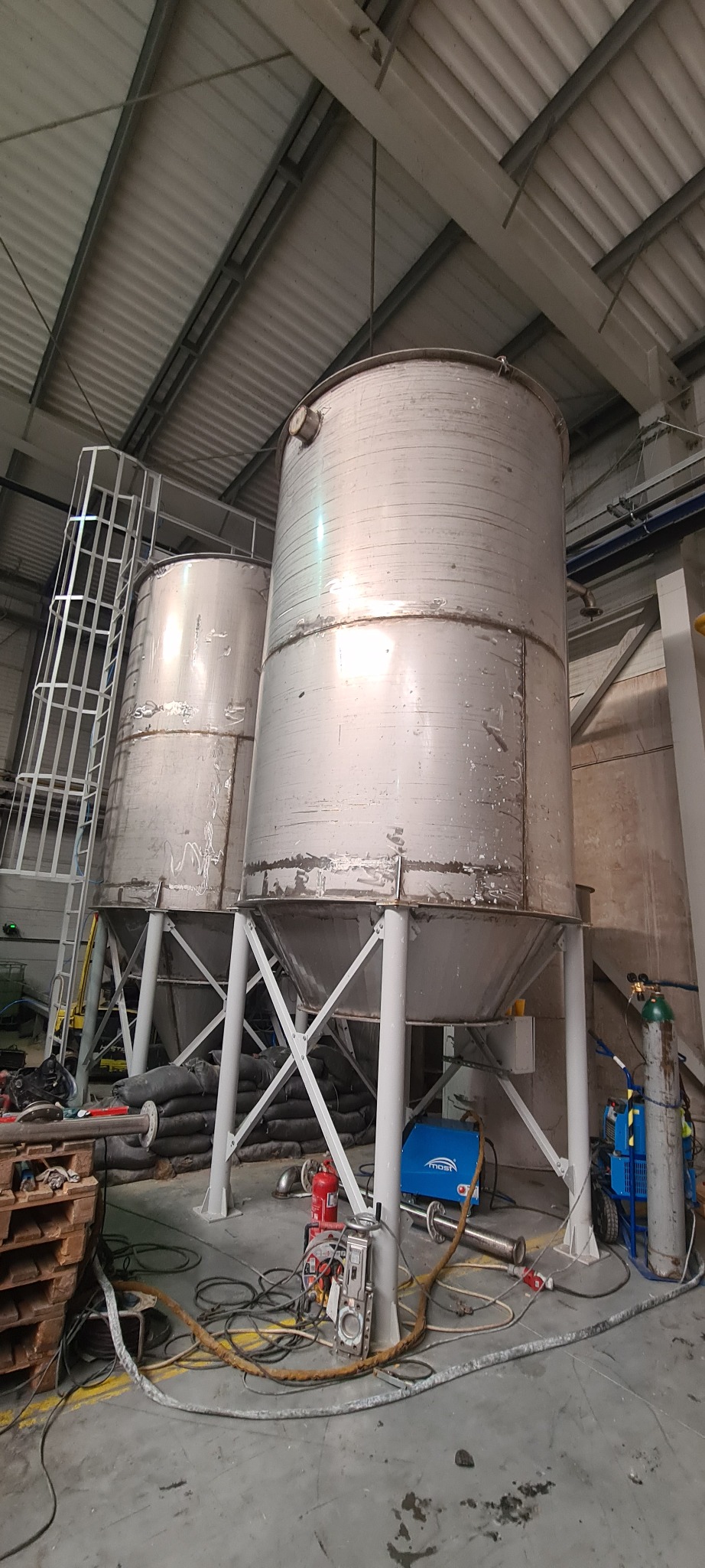 Dwa duże, metalowe zbiorniki przemysłowe na stalowych podporach w hali produkcyjnej, widoczna spawarka i butle z gazem, sugerujące prace spawalnicze.