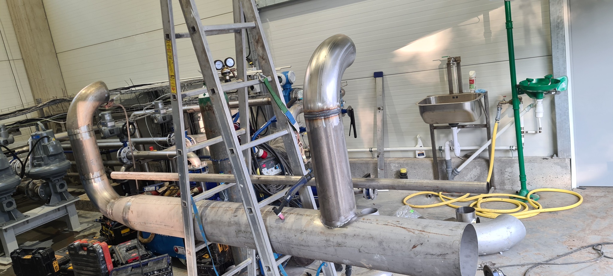 Spawanie rur ze stali nierdzewnej w hali produkcyjnej, widoczne spoiny, drabina, narzędzia i instalacje.