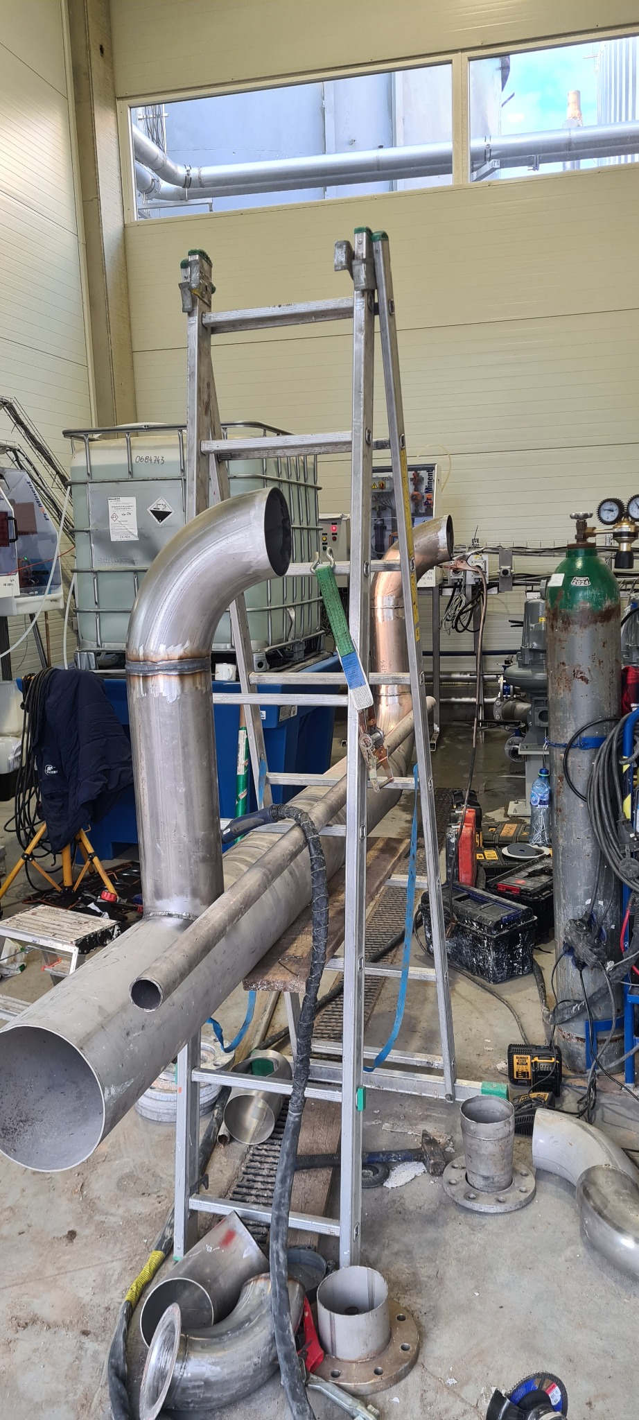 Wewnętrzny widok hali produkcyjnej z fragmentami spawanych rur ze stali nierdzewnej na drabinie, widoczne butle z gazem technicznym i zbiornik IBC w tle.