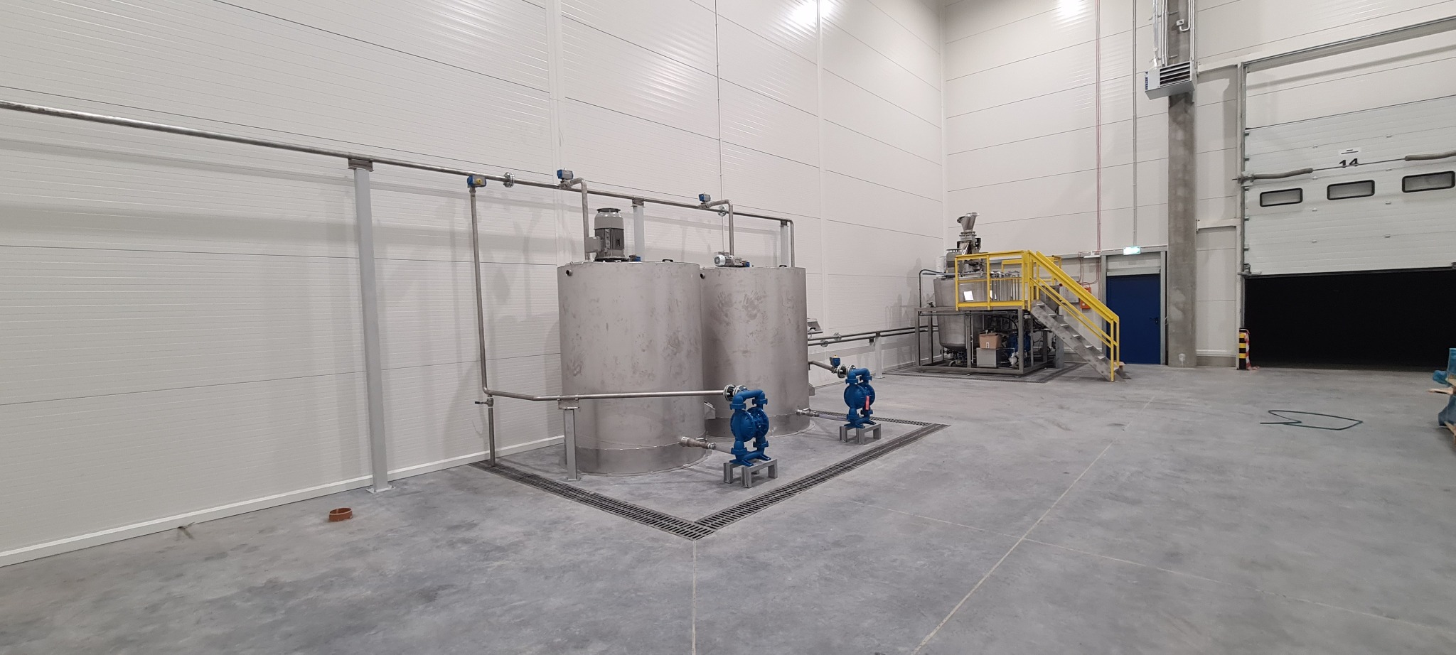 Dwa stalowe zbiorniki przemysłowe z niebieskimi pompami membranowymi w hali produkcyjnej z kratkami ściekowymi i instalacją rurową.