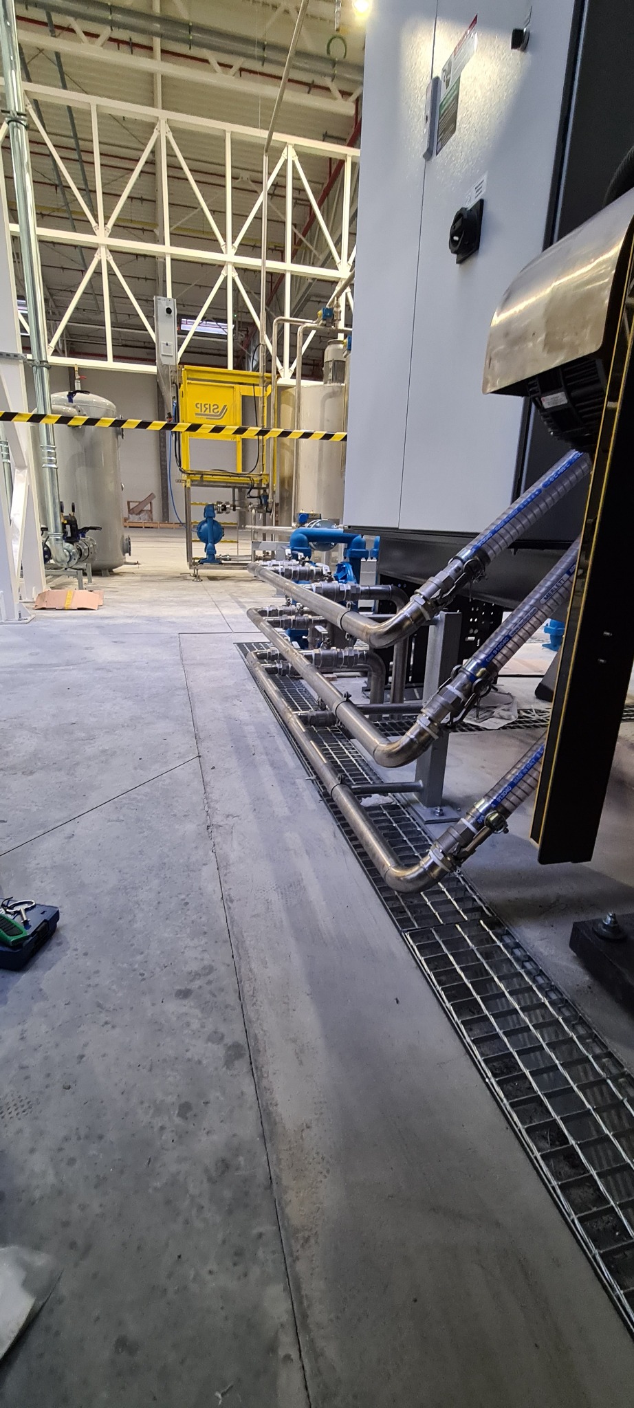 Instalacja przemysłowa z rurami ze stali nierdzewnej, widoczne połączenia i zawory, kratka ściekowa w posadzce, w tle zbiorniki i konstrukcja hali.