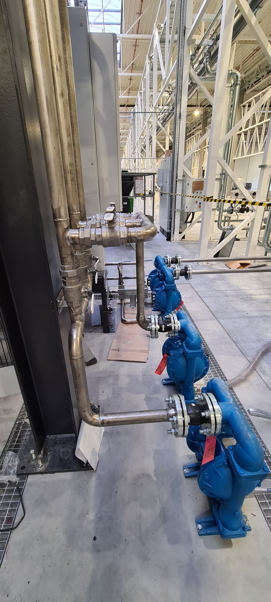 Rząd trzech niebieskich pomp przemysłowych połączonych rurami ze stali nierdzewnej w hali produkcyjnej.