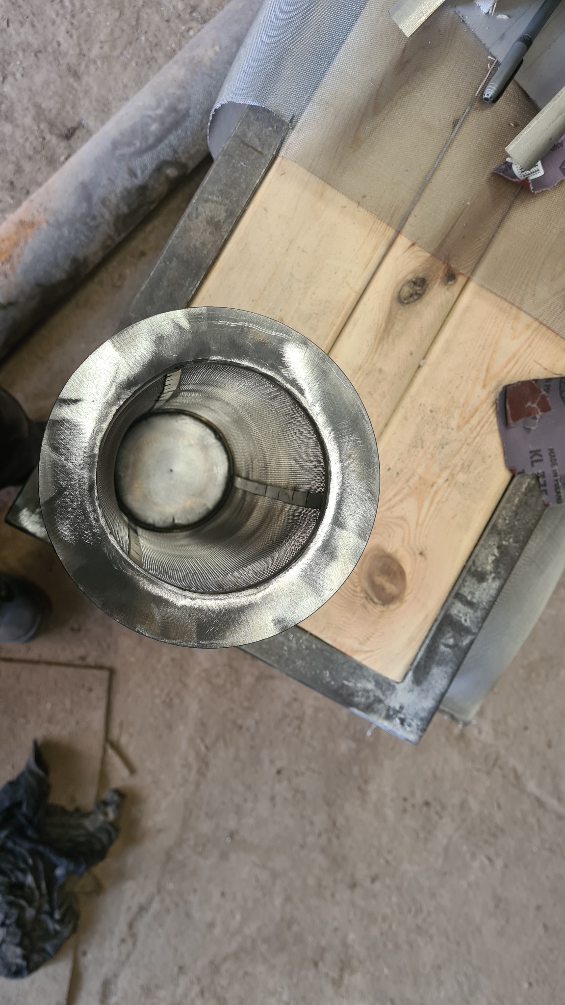 Metalowy cylinder z siatką w środku, widoczny z góry, leżący na drewnianym blacie z metalową ramą. Widoczne ślady obróbki i niedoskonałości powierzchni.