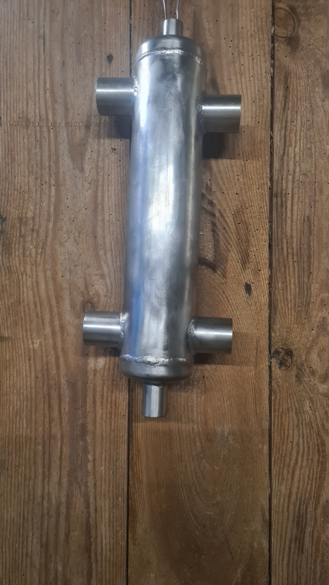 Surowy, spawany wymiennik ciepła ze stali nierdzewnej z czterema króćcami, zawieszony na tle drewnianej ściany.