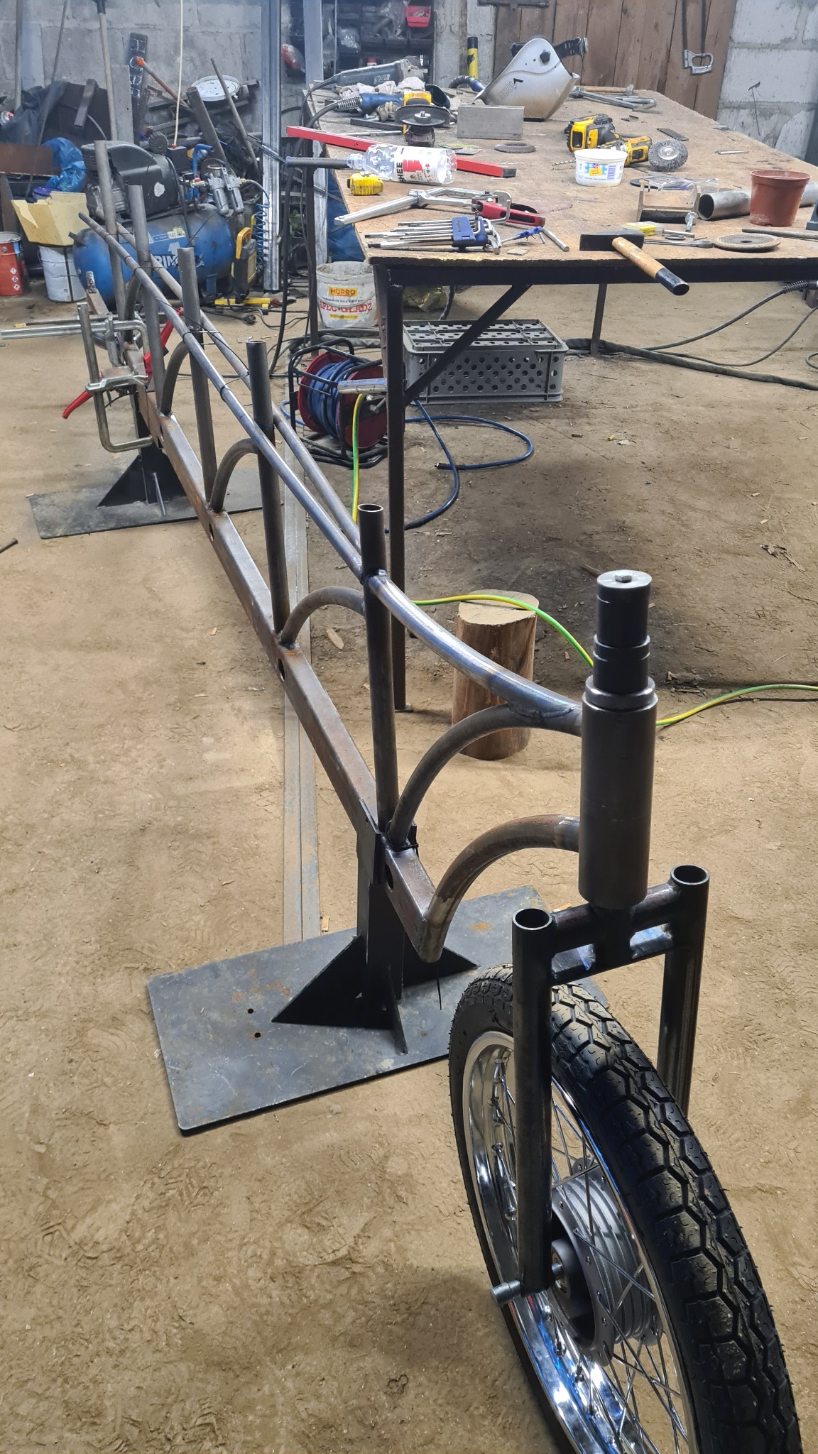 Metalowa konstrukcja w warsztacie, przypominająca ramę wózka lub roweru, spawana na metalowych podstawach, z widocznym kołem i narzędziami w tle.