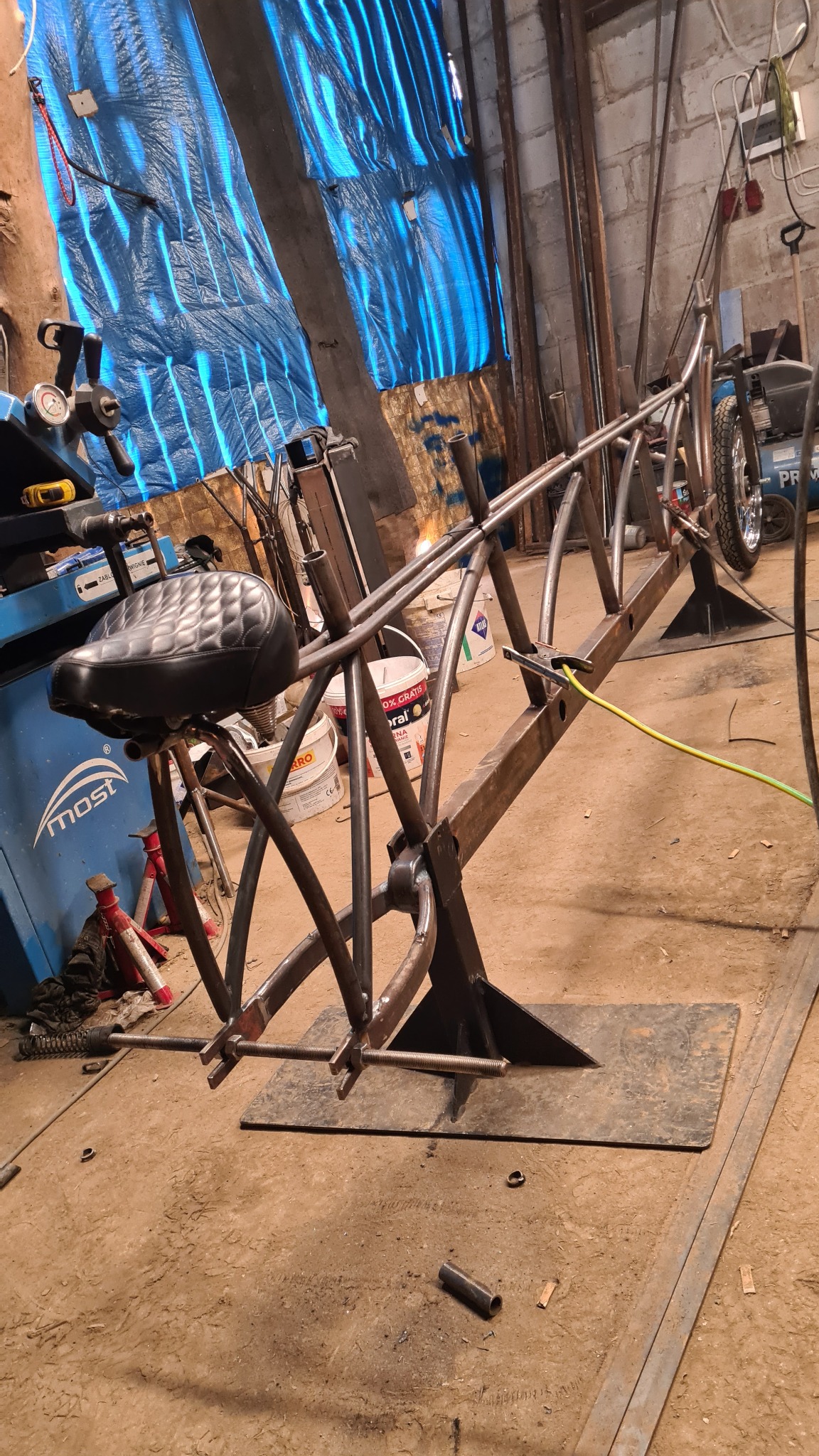 Rama roweru typu chopper w trakcie budowy, spawana z metalowych rur, z zamontowanym skórzanym siodełkiem, widoczne narzędzia i warsztatowe tło.