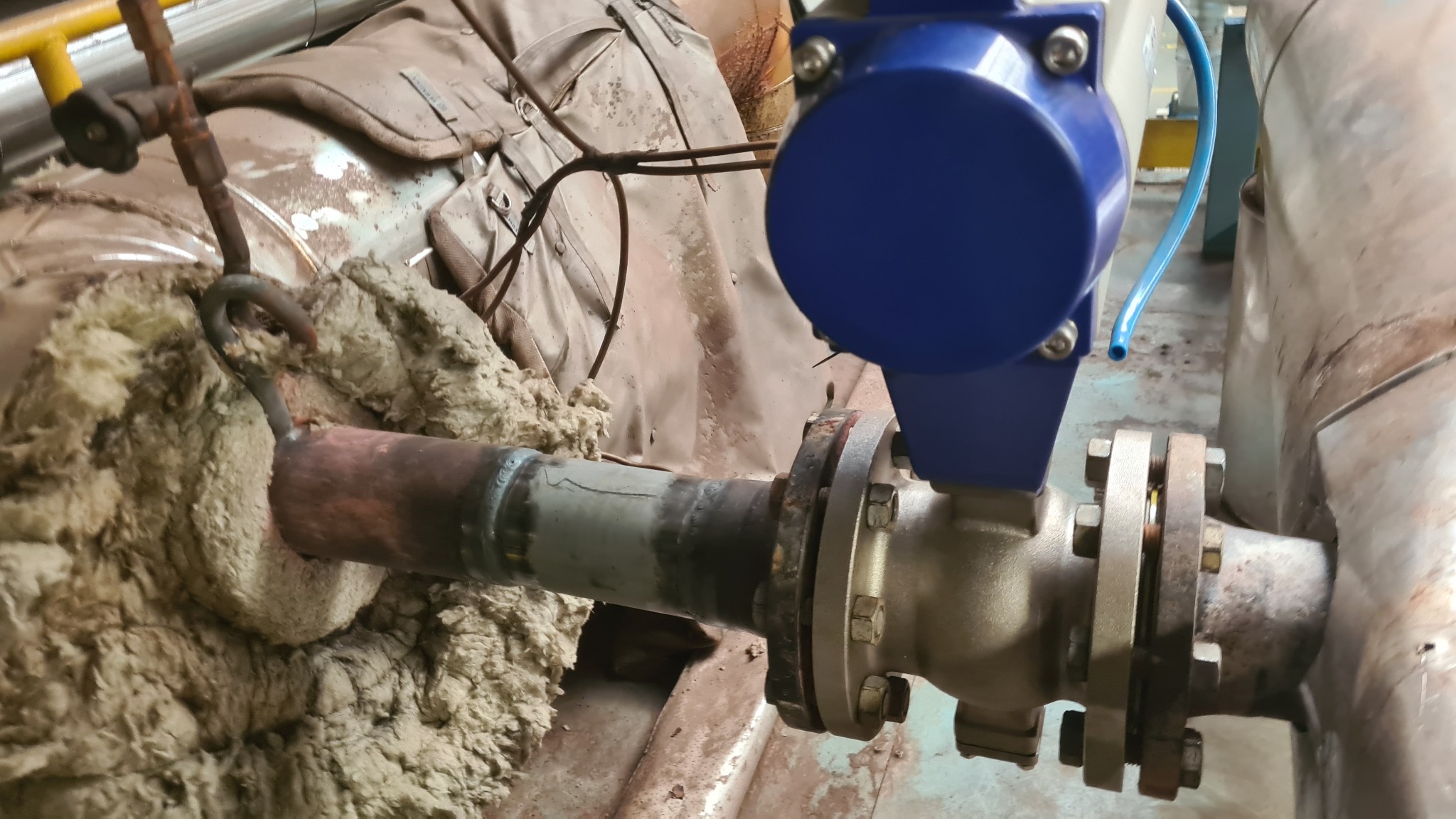 Zespawane rury z kołnierzem i niebieskim zaworem, częściowo owinięte izolacją, widoczne ślady spawania.