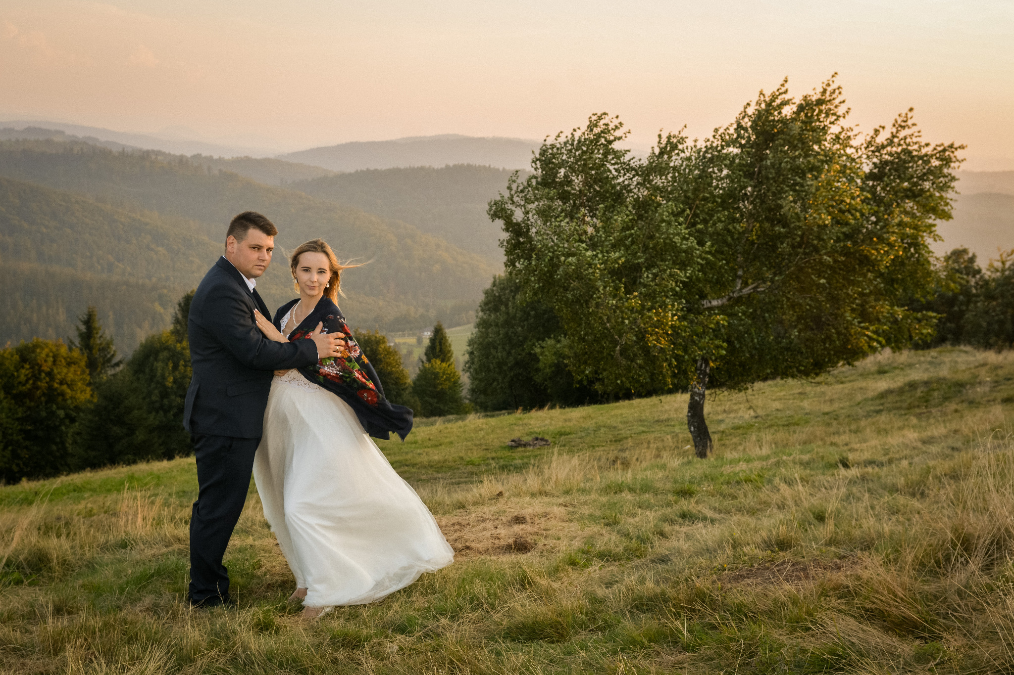 Para młoda w objęciach na tle górskiego krajobrazu o zachodzie słońca, panna młoda w białej sukni i kwiecistym szalu.