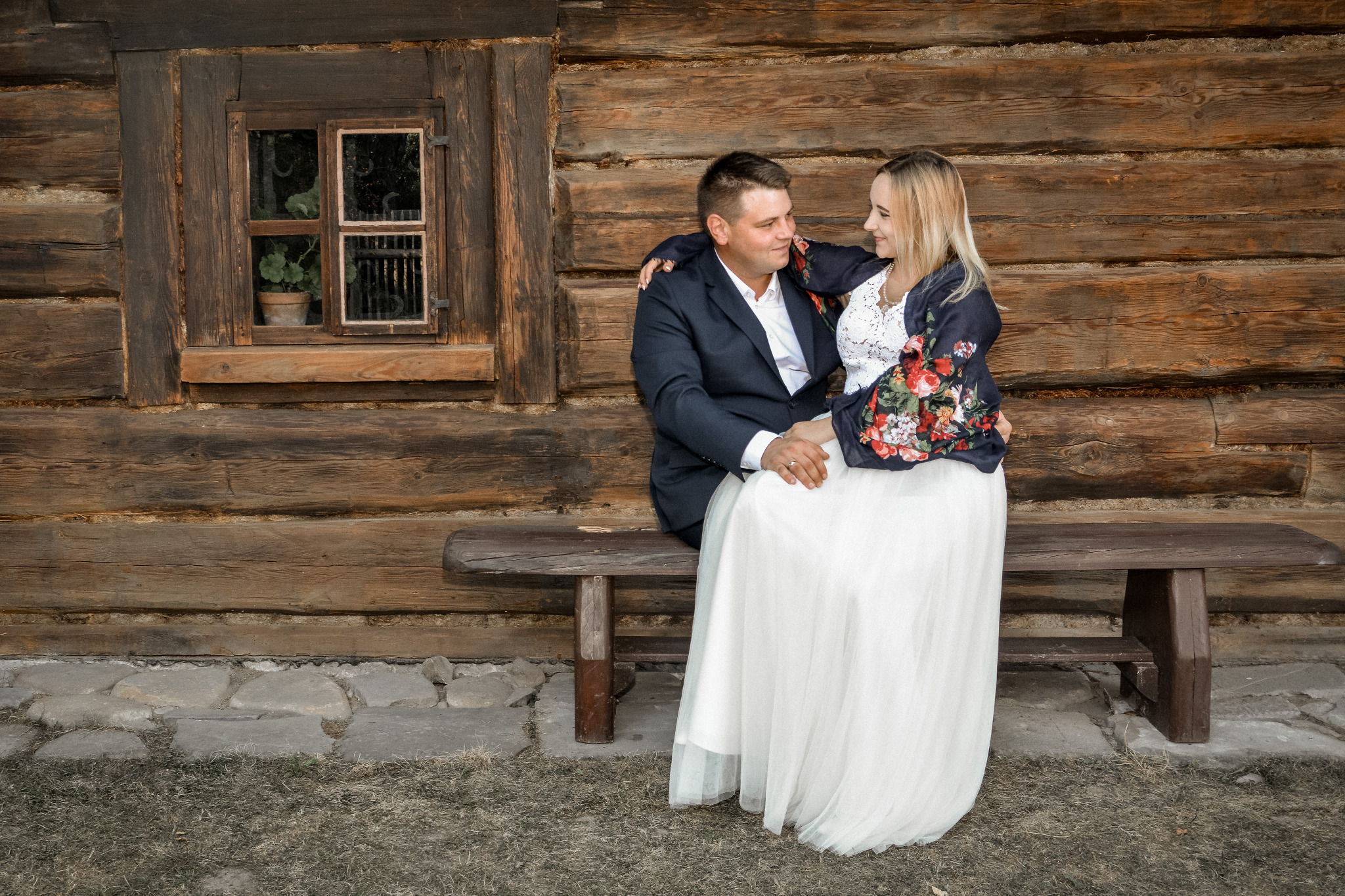 Para młoda siedzi na ławce przed drewnianym domem, panna młoda w białej sukni i kwiecistej narzutce, pan młody w granatowym garniturze, oboje patrzą na siebie z uśmiechem.