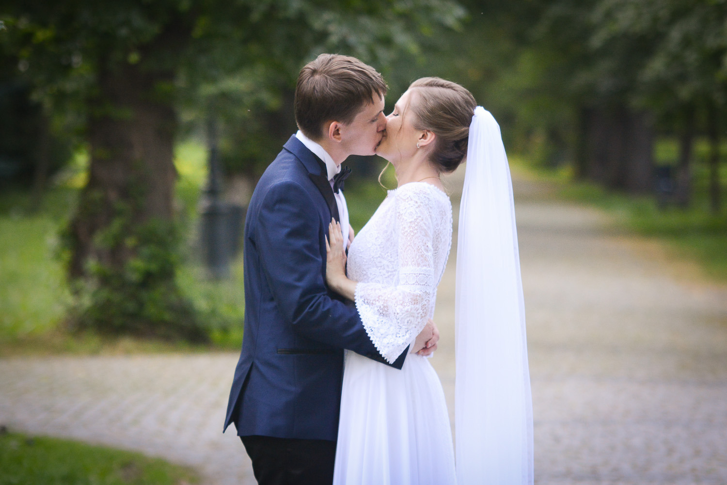 Pocałunek pary młodej w plenerze, panna młoda w koronkowej sukni z welonem, pan młody w granatowym smokingu z muszką, parkowe tło.
