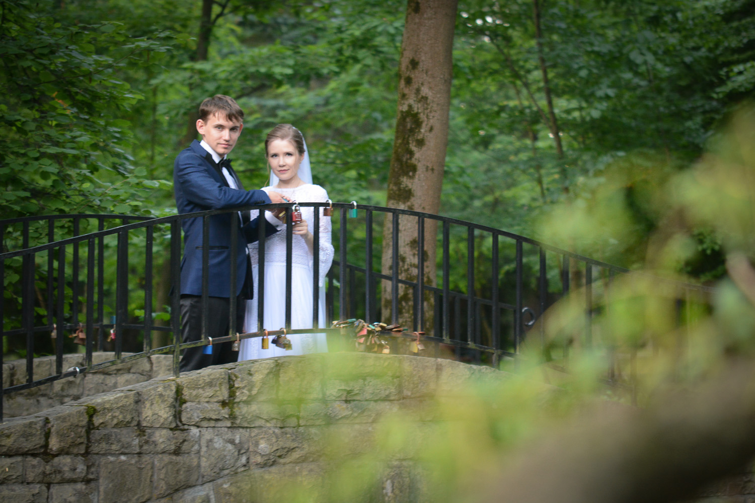 Para młoda w strojach ślubnych stoi na kamiennym moście z czarną metalową barierką, do której przyczepione są kłódki, w tle zielony las.