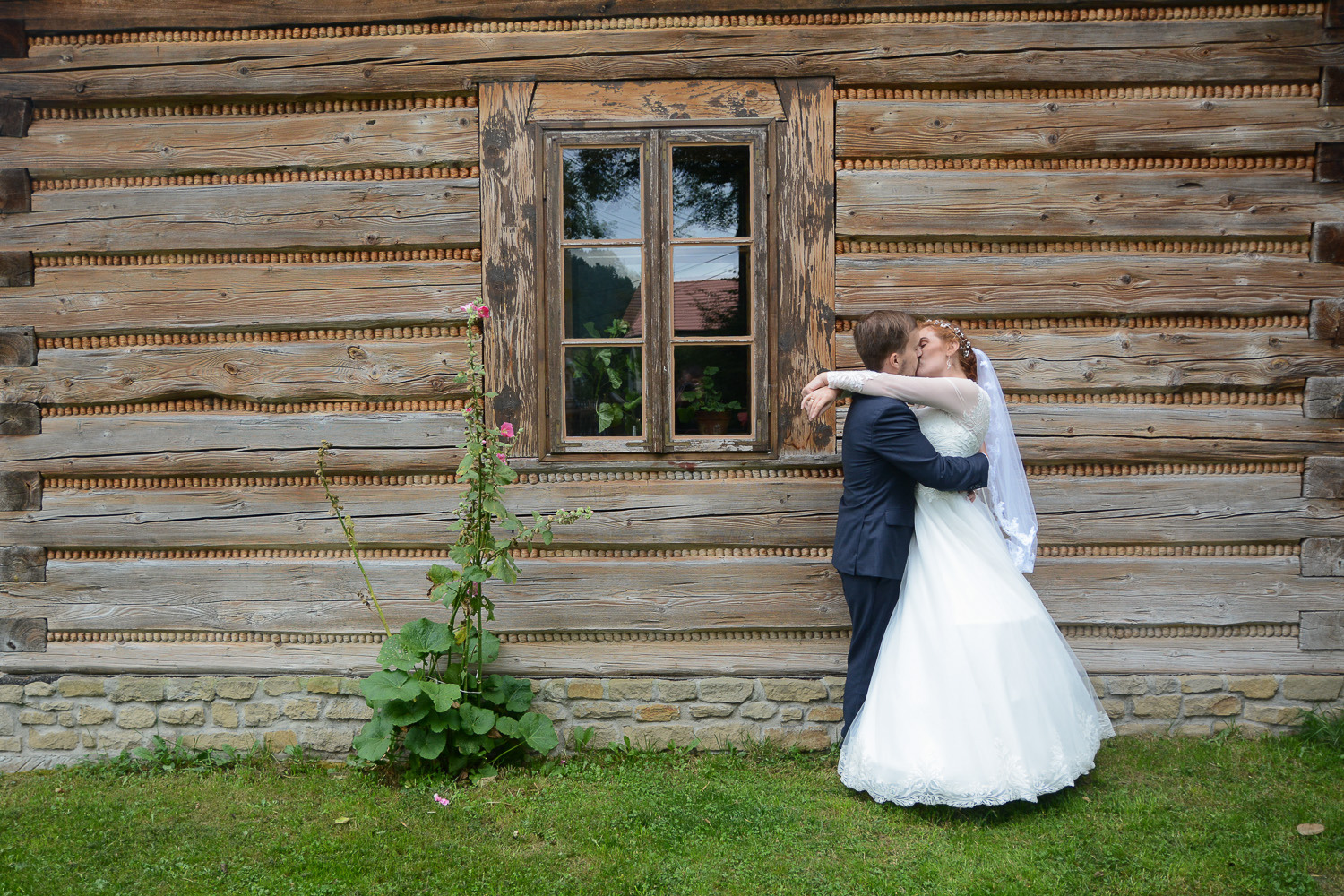 Pocałunek nowożeńców na tle drewnianej ściany z oknem, w tle widoczny fragment czerwonego dachu.